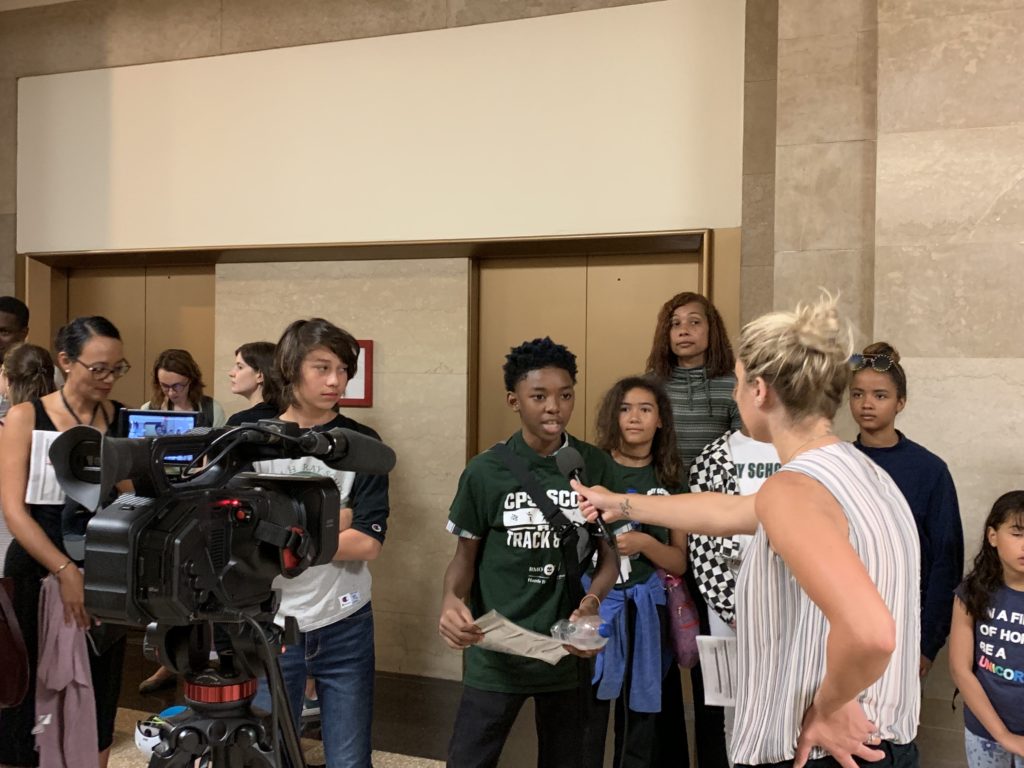 A diverse group of students is being interviewed by a female reporter in front of elevators. A video camera captures the scene.