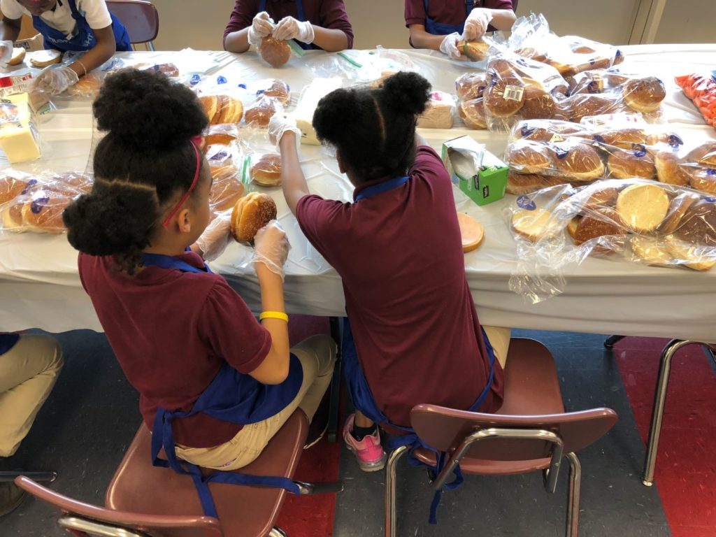 Two children in burgundy shirts and blue aprons sit at a table assembling sandwiches with bread. They wear gloves, showing teamwork and focus.