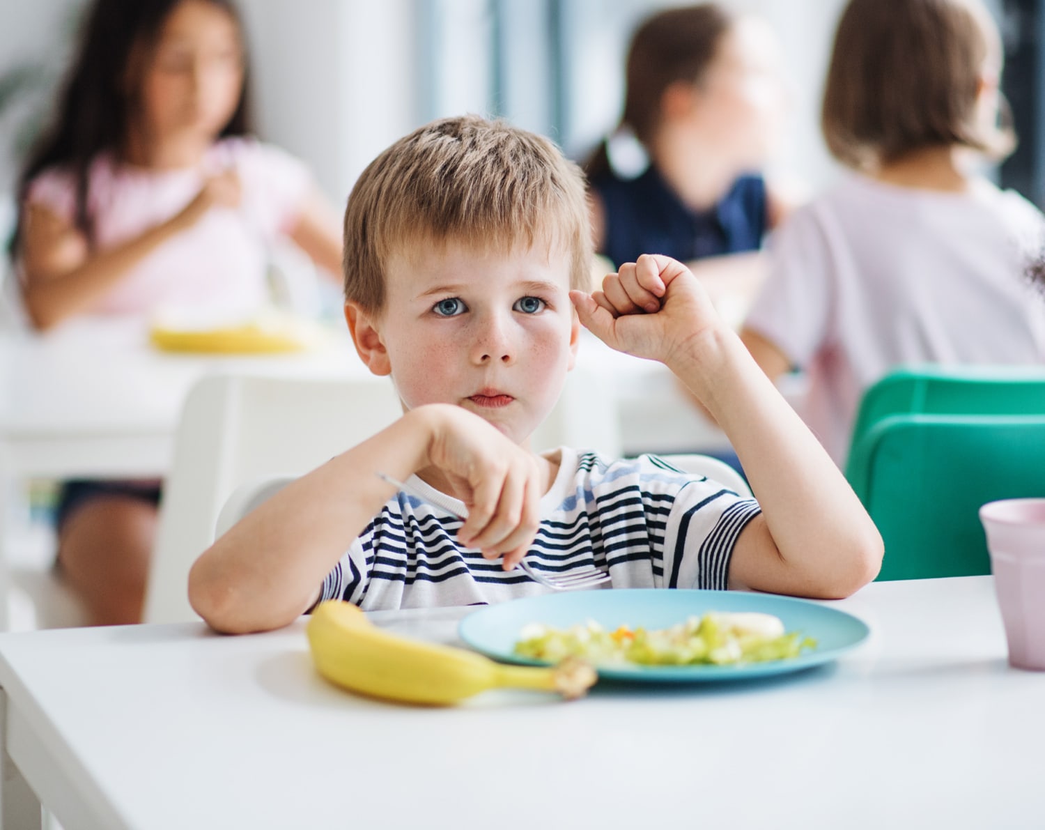 A young boy in a striped shirt sits thoughtfully at a table with a plate of salad and a banana. Other children are blurred in the background.