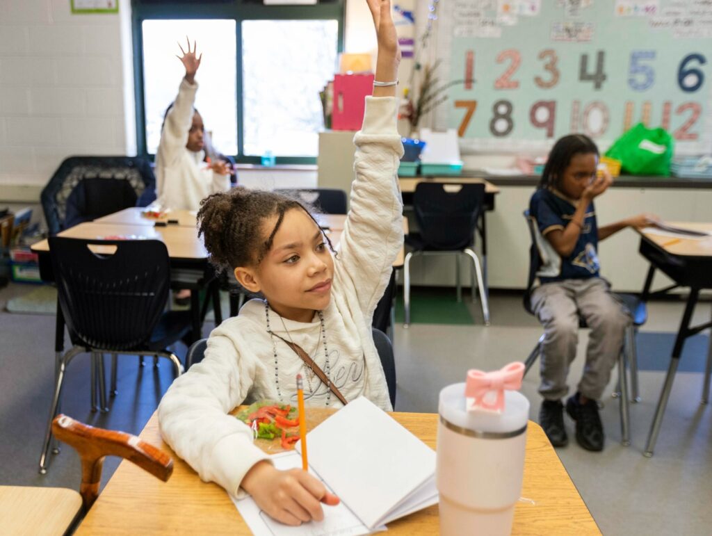 A young girl in a classroom eagerly raises her hand, holding a pencil over a notebook. Other children sit at desks in the background. The mood is focused and engaged.