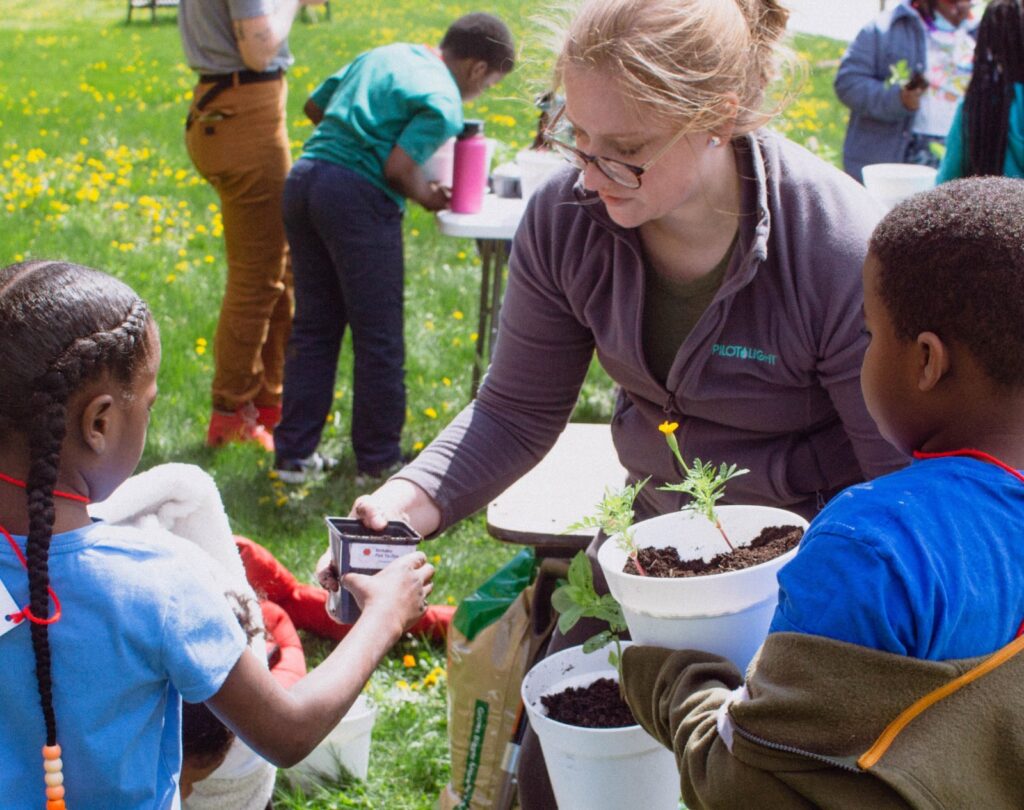 A woman assists children in planting seeds outdoors. She passes a small container to a girl. Nearby, a boy holds a potted plant. The scene is lively and educational.
