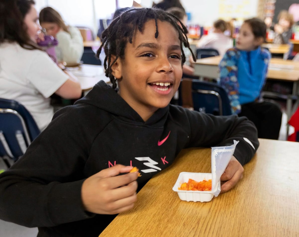 A smiling child with braided hair sits at a classroom desk, wearing a black hoodie. He holds a container of orange snacks, surrounded by other students.