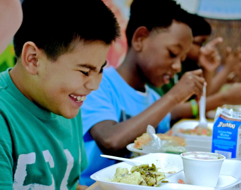 Two children sit at a table, smiling and eating lunch. The child in front wears a green shirt, while the one behind wears blue, creating a joyful atmosphere.